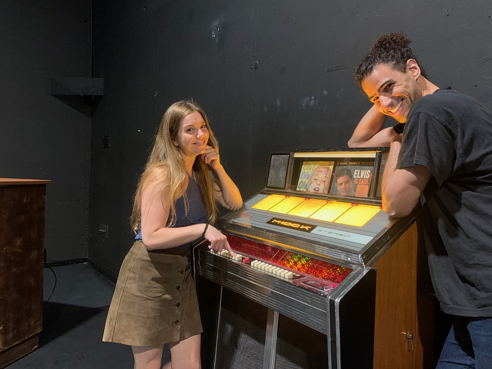 Emma Jane Howely und Gustavo de Oliveira Leite im Bühnenbild von „Gradually, Then Suddenly” mit der magischen Jukebox. Das vorliegende Modell „Harting M100“ verfügt übrigens über die rote Hit-Automatik-Taste. Der Gast musste die Hits nicht einzeln wählen, sondern sie konnten  mittels der Hit-Automatik-Taste entsprechend des Kredits alle auf einmal abgespielt werden.